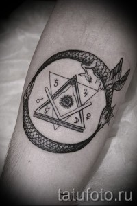 Фото тату Уробороса, окруженного масонскими символами, выполненного в технике blackwork на предплечье. Детализированный рисунок.