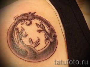 Фото тату: изображение Уробороса – змеи, кусающей свой хвост, в котором тело змеи переходит в фигуру волка, выполненное в черно-серых тонах на плече.