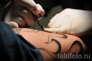 Фото процесса татуировки: крупный план тату-машинки, работающей на коже клиента. Видны капли крови и чернила, а также руки мастера в перчатках.