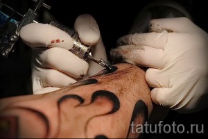 Фото процесса татуировки: мастер в перчатках наносит рисунок на кожу клиента с помощью тату-машинки. Видны капли крови и чернила.