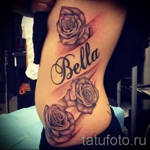 Фото тату: две розы и надпись "Bella" на женском боку. Рисунок выполнен в реалистичном стиле с использованием черно-серых тонов.