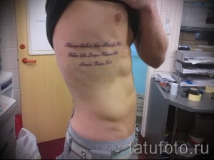 Фото тату: длинная цитата из Библии, выполненная черными чернилами на боку мужчины. Текст расположен вертикально вдоль ребер.