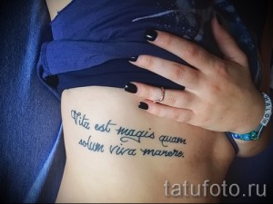 Фото тату: изящная надпись на латыни 'Vita est magis quam solum viva manere' на боку девушки, выполненная черными чернилами.
