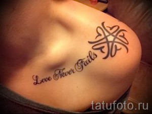 Фото тату: небольшая татуировка на ключице, состоящая из надписи "Love Never Fails" и символического изображения. Изящные линии и темный цвет создают эффект элегантности.