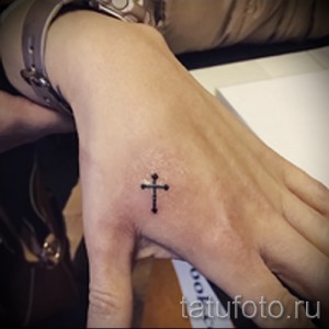 Фото тату: маленький черный крест, выполненный в минималистичном стиле, на тыльной стороне ладони. Простой и лаконичный дизайн.
