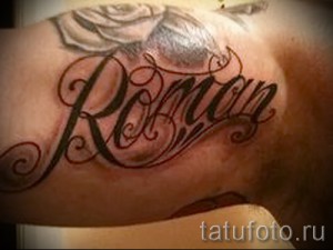 Фото татуировки с именем 'Roman' и изображением розы на плече. Черно-красный рисунок татуировки, выполненный в реалистичном стиле.