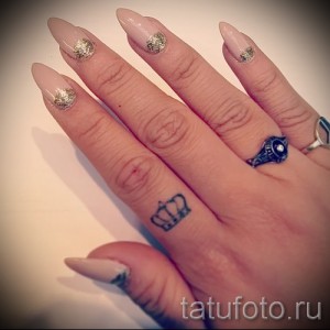 Фотография маленькой татуировки в виде короны на безымянном пальце руки. Черно-белый рисунок татуировки выполнен тонкими линиями.