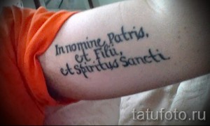 Фото татуировки с латинской фразой 'In nomine Patris, et Filii, et Spiritus Sancti' на внутренней стороне предплечья.