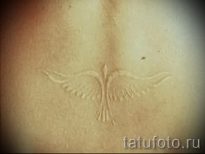 Фотография белой татуировки на спине, изображающей птицу с расправленными крыльями. Изящный рисунок татуировки выполнен в технике white ink.