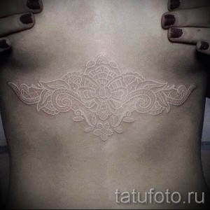 Фото белой татуировки мандалы на груди женщины. Сложный геометрический узор выглядит изящно и загадочно.