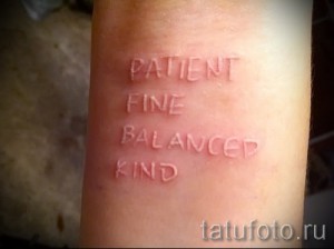 Фото белой татуировки на предплечье с надписью 'PATIENT FINE BALANCED KIND'. Минималистичный дизайн, светлые тона, нежный рисунок татуировки.