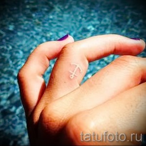 Фотография белой татуировки в виде якоря на пальце руки. Минималистичный и стильный дизайн.