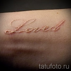 Фотография белой татуировки надписи "Loved" на запястье. Простой и лаконичный рисунок, выполненный тонкими линиями.