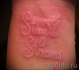 Фото белой татуировки с надписью 'Stay Strong' и изображением крыльев на запястье. Вдохновляющий рисунок, выполненный каллиграфическим шрифтом.