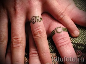Фото тату на пальцах: две руки с татуировками инициалов 'M' и 'C', обвитых зелеными листьями и стеблями. Нежный и романтичный дизайн.