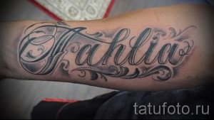 Фото тату с именем 'Aahlia', выполненным в стиле каллиграфии с добавлением черных узоров и теней на предплечье.