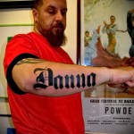 Фото тату с именем 'Donna' на предплечье мужчины, выполненное в готическом стиле черными чернилами. Мужчина с бородой смотрит в камеру.