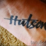 Фото тату с надписью 'Hudson' на стопе. Черный шрифт, выполненный в стиле леттеринга. Минималистичный дизайн.