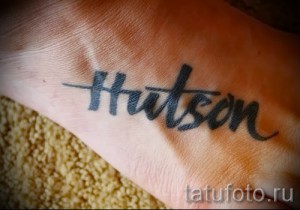 Фото тату с надписью 'Hudson' на стопе. Черный шрифт, выполненный в стиле леттеринга. Минималистичный дизайн.
