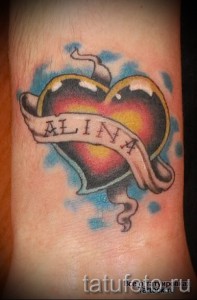Фото тату сердца с именем «Alina» на руке. Татуировка выполнена в стиле олдскул с использованием ярких цветов: красного, желтого, голубого и черного.