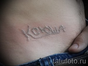 Фото старой, выцветшей татуировки с надписью 'Коля' на животе, с нечеткими контурами и следами времени.