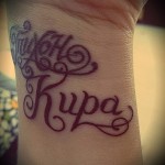 Фото тату с именем 'Kupa', выполненным фиолетовым шрифтом с декоративными узорами на запястье, создающим нежный и романтичный образ.