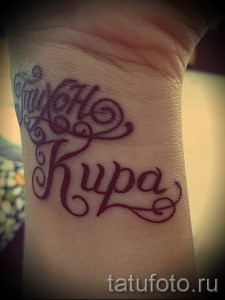 Фото тату с именем 'Kupa', выполненным фиолетовым шрифтом с декоративными узорами на запястье, создающим нежный и романтичный образ.