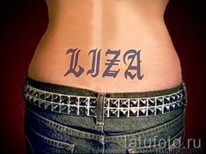 Фото тату с именем 'Liza', выполненным готическим шрифтом на пояснице девушки, с акцентом на стильный ремень с шипами.