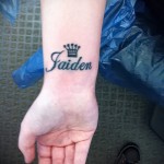 Фото татуировки с именем 'Faiden' и изображением короны. Черный контур, минималистичный дизайн, нанесена на запястье.