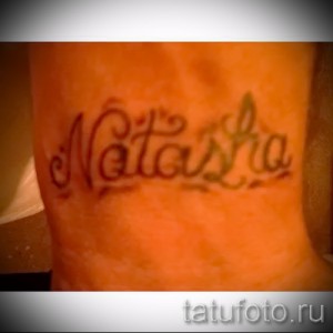Фото тату с именем 'Natasha', выполненное каллиграфическим шрифтом с декоративными завитушками на лодыжке. Черно-серая татуировка.