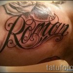 Фото тату надписи 'Roman' на шее. Черные чернила, каллиграфический шрифт, декоративные завитки и узоры. Элегантный и привлекающий внимание дизайн.