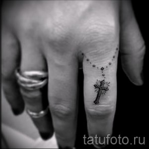 Черно-белое фото татуировки на пальце: четки с крестом. Детализированный рисунок, религиозная символика, минималистичный стиль.