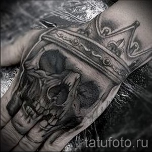 Фото тату большой короны, наложенной на череп, на руке. Детализированный рисунок татуировки в стиле реализм.