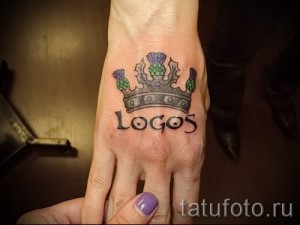 Фото тату короны с чертополохом и надписью "Logos" на руке. Цветной рисунок татуировки с использованием синих, зеленых и серых оттенков.
