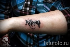 Фото тату с именем 'Alyssa', выполненным черными чернилами с добавлением красных брызг краски, расположенным на предплечье.