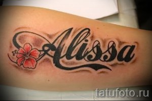 Фото тату на предплечье: имя "Alissa" с изображением цветка гибискуса, выполненное в ярких цветах, на женской руке.