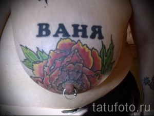Фото тату на животе: большой красный пион с зелеными листьями и надпись 'Ваня' сверху. Цветная татуировка, реалистичный стиль, женственный дизайн.