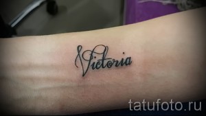 Фото татуировки с именем "Victoria", выполненным изящным каллиграфическим шрифтом на запястье. Татуировка выполнена в черных тонах.