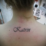 Фото тату надписи 'Katrin' изящным шрифтом на спине, чуть ниже шеи. Женственная татуировка, выполненная в черных тонах.