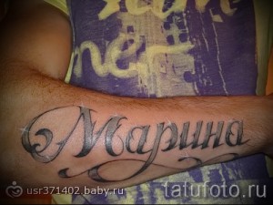 Фото тату надписи 'Марина' на предплечье, выполненной в каллиграфическом стиле с использованием черных чернил и декоративных завитков.