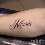 Фото тату надписи 'Maria' на женском предплечье, выполненной в каллиграфическом стиле с использованием коричневых чернил и тонких линий.