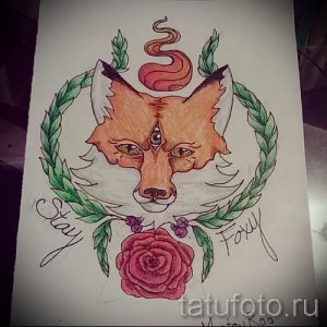 Фото татуировки лисы с третьим глазом на лбу, окруженной лавровым венком и розовыми цветами. Рисунок выполнен в ярких, насыщенных цветах.