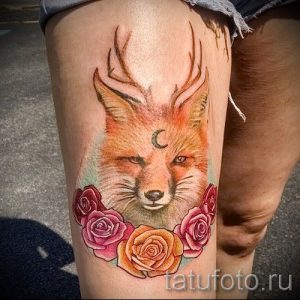 Фото татуировки лисы с рогами и луной на бедре. Рисунок выполнен в оранжевых и розовых тонах, с добавлением цветочных элементов.