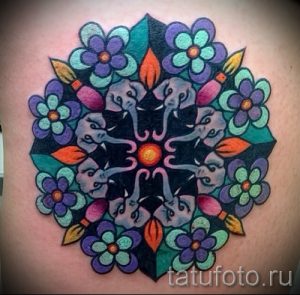 Фото тату мандалы с изображением слонов и цветов. Цветная татуировка с использованием ярких оттенков синего, зеленого, оранжевого и фиолетового, расположенная на плече.