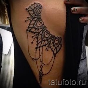 Фотография коричневой татуировки совы, стилизованной под мандалу, на бедре девушки. Детализированный рисунок с геометрическими узорами и точками.
