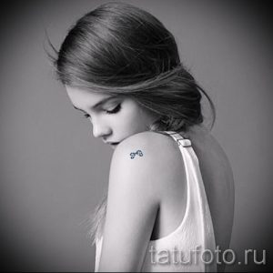 Фото тату с изображением маленького бантика на ключице девушки. Минималистичный и элегантный рисунок татуировки в черном цвете.