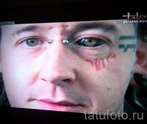 Фото тату на лице мужчины: красные линии, напоминающие царапины, и черные символы на виске. Экстремальный рисунок татуировки.