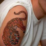 Фото тату на плече мужчины. Изображен тигр в прыжке, выполненный в ярких оранжевых и черных тонах.
