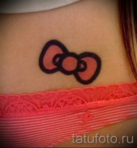 Фото тату на шее: изображение бантика Hello Kitty, выполненное в нежном розовом цвете с черным контуром.