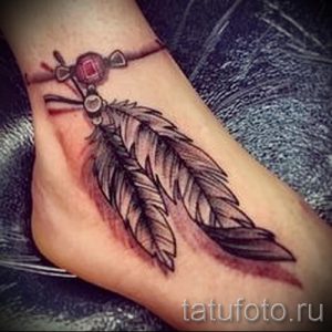 Фото тату: красочное перо и индейское украшение с красным камнем на стопе. Детализированный рисунок татуировки в ярких тонах.
